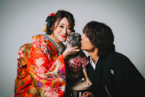 和装を着た女性と男性が、間にいる犬にキスをしているシーン。女性は華やかな着物を着用。