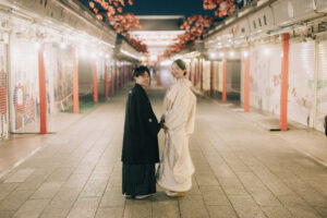 和装を着た二人が手をつないでいる様子。背景には夜の街並みと花の装飾が見える。