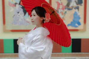 赤い傘を持った和装の女性が、伝統的な背景の前で静かに立っている。白い着物と花の髪飾りが特徴的。