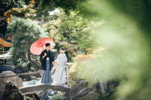 和装の男女が赤い傘を持ちながら橋の上で立っている様子。背景には緑豊かな自然が広がっている。