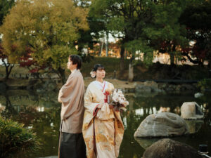 和装を着た男女が池の近くで立っている。女性は花束を持ち、男性は背を向けている。