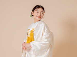 白い和装を着た女性が微笑みながら立っている。髪には緑の飾りがあり、柔らかな背景でポーズを取っている。