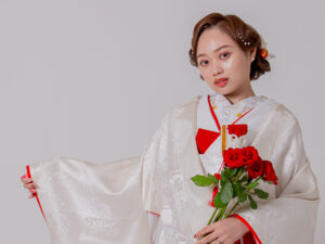 白い着物を着た女性が赤いバラの花束を持っている。彼女の髪には花のアクセサリーが飾られている。