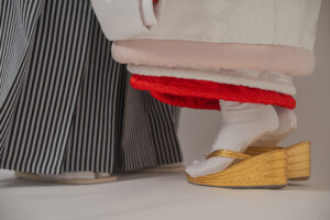 白い和装を着た人物の足元と、ストライプの着物を着た人物の足元が映る。和装の下に履かれた金色の草履が特徴的。