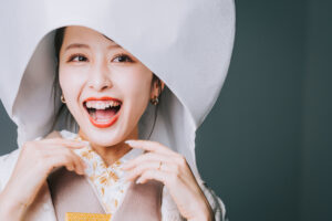 白い帽子をかぶった女性が笑顔でポーズをとっている。彼女は和装を着ており、明るい表情を見せている。