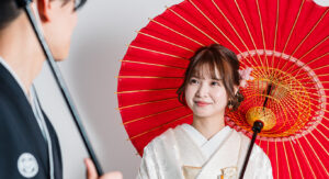 赤い傘を持つ和装の女性が微笑んでいる様子。彼女は白い着物を着用し、髪に花のアクセサリーをつけている。