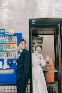 新郎新婦が公衆電話ボックスの前で笑顔を見せている。新婦は白いドレスを着用し、新郎はダークカラーのスーツを着ている。