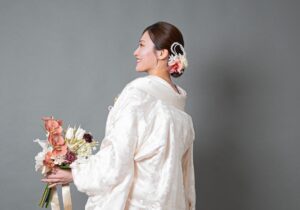 白い和装を着た女性が花束を持ち、後ろを向いている。髪には花のアクセサリーが付いている。