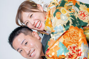 和装を着た女性が男性の背中に乗って笑顔を見せているシーン。鮮やかな花柄の着物が特徴的。