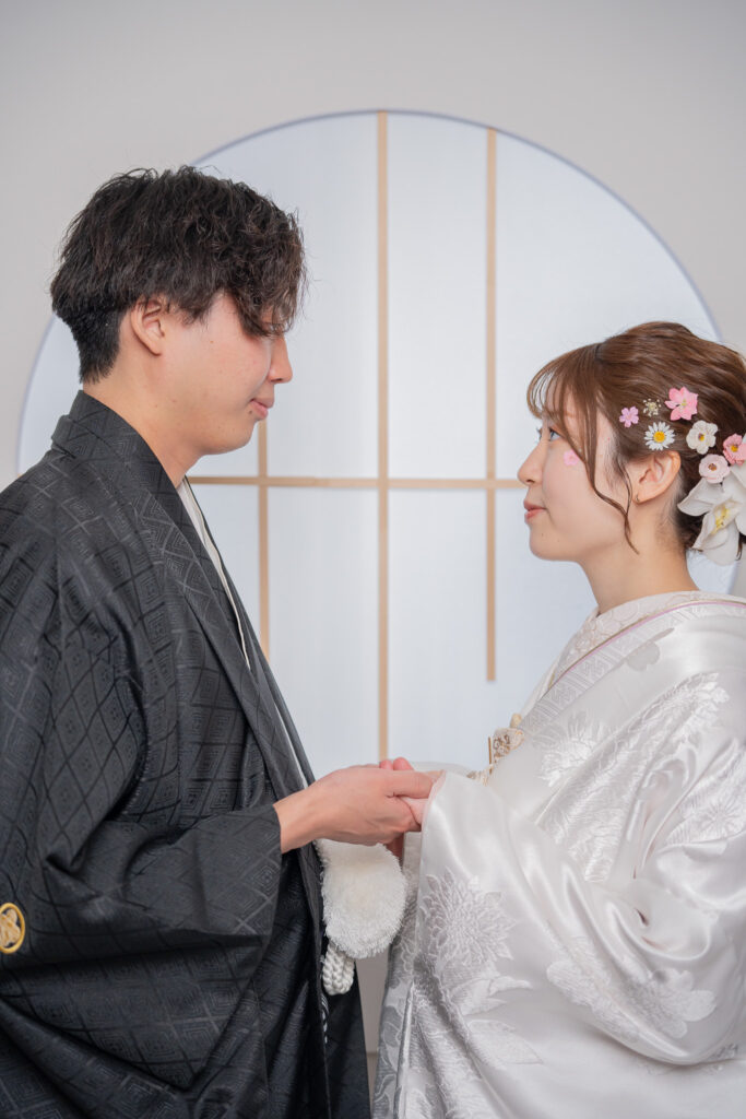 浅草店限定の窓枠背景で手と手を取り合う白無垢姿の花嫁と光沢のある黒紋付き姿の新郎のショット