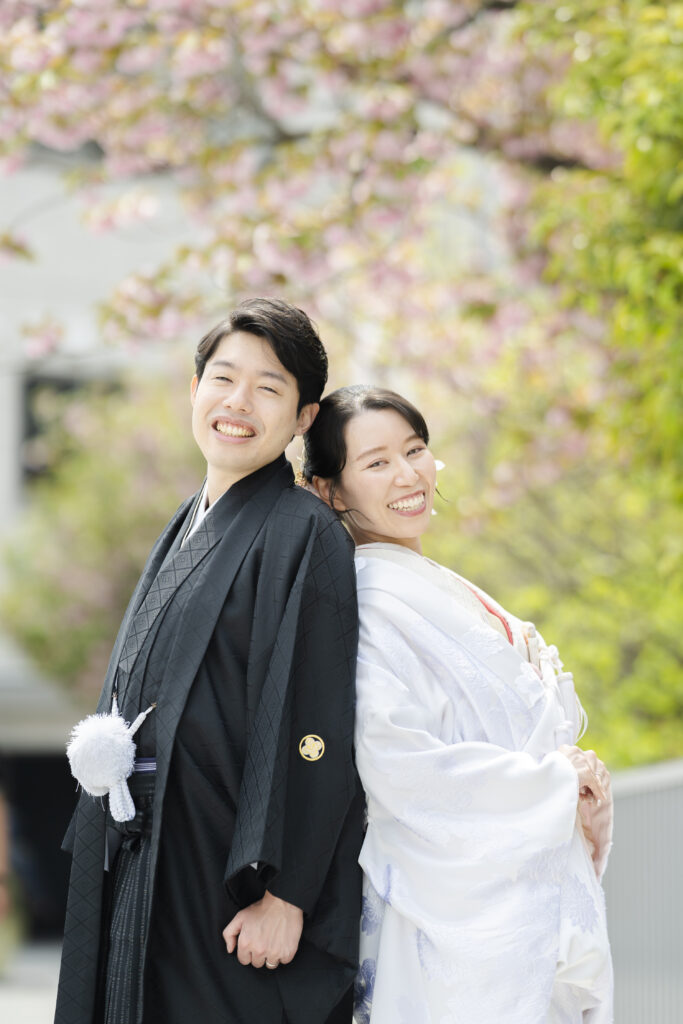 桜の木の下で和装前撮りを楽しむ白無垢姿の花嫁と黒紋付羽織袴の新郎様のショット