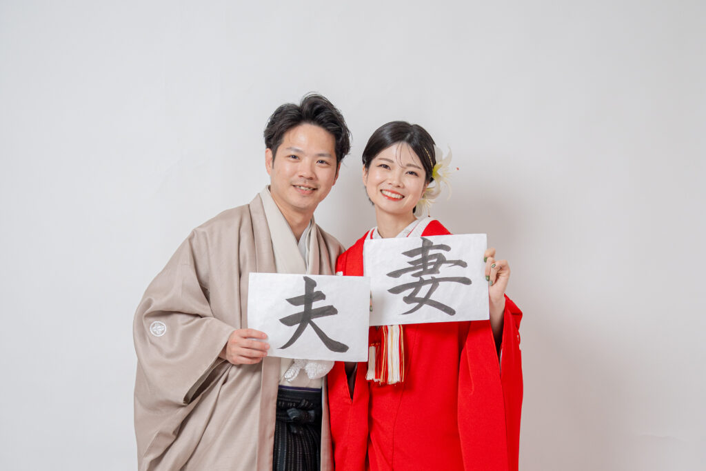 2ショット。手書き（習字）「夫」「妻」をもって笑顔で。色打掛（赤）。横向き