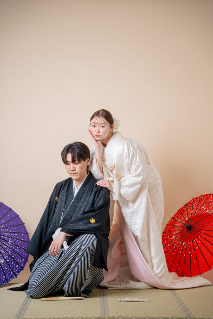 畳で新郎の肩に手を置く白無垢新婦。番傘。