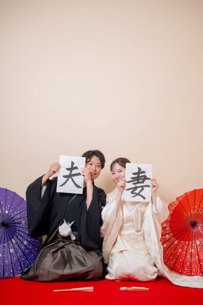 和装の2人が正座で「夫」「妻」の習字から顔をのぞかせる新婚らしい2ショット。紋付き袴、白無垢、番傘