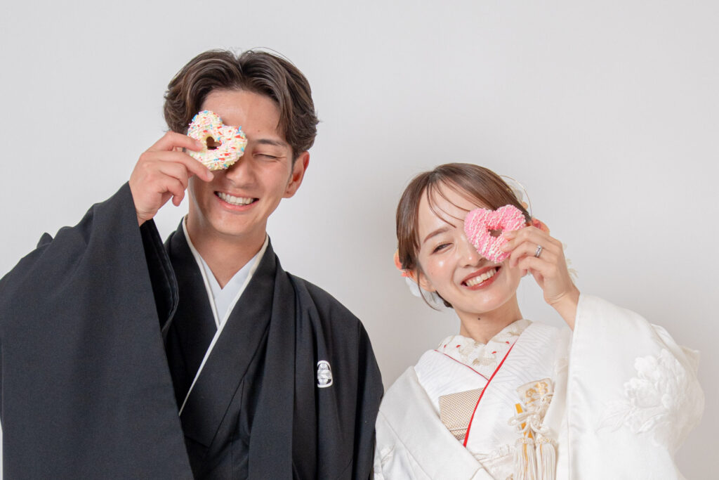 和装の新郎新婦がドーナツのおもちゃを片手に目を閉じ笑顔のショット。白無垢