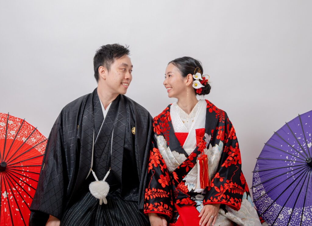 和装の新郎新婦が笑顔で見つめあう、座り2ショット。紋付き袴、黒字に白と赤の模様が色鮮やかな打掛。番傘