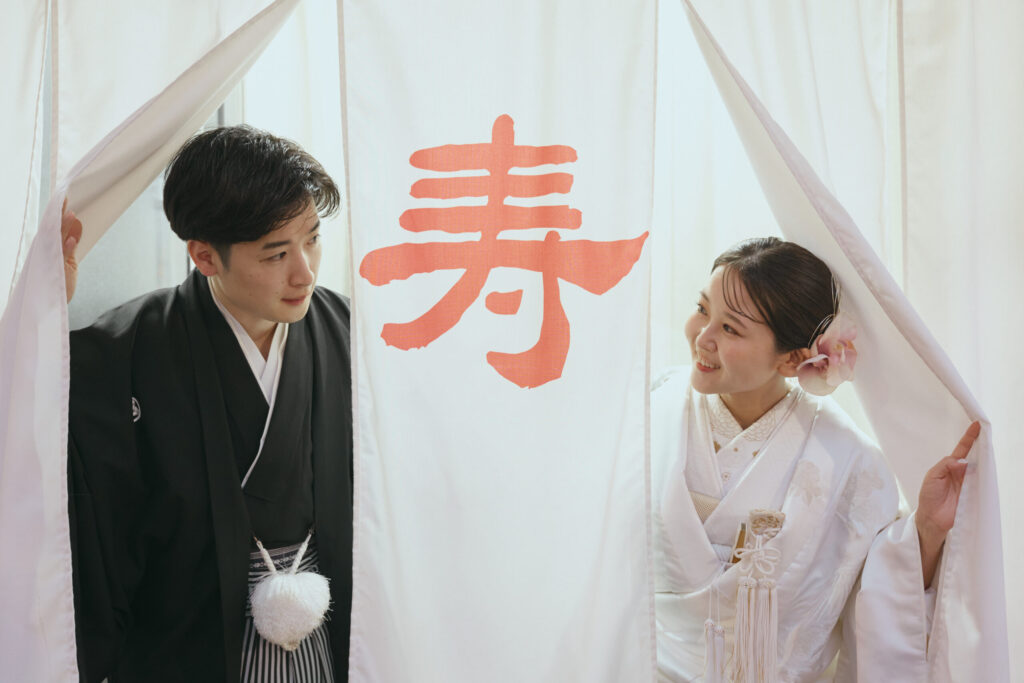 和装の新郎新婦が暖簾から顔をのぞかせたかわいらしい2ショット。紋付き袴、白無垢
