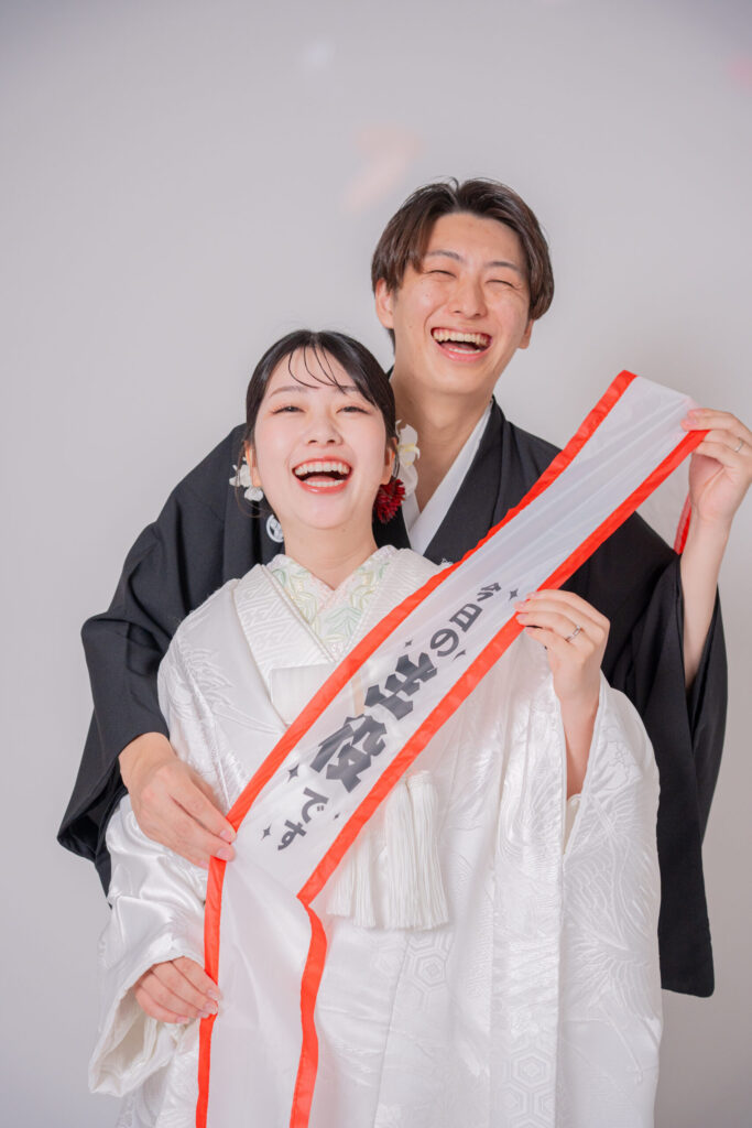 和装の新郎新婦が自然体笑顔の2ショット。主役たすき、紋付き袴、白無垢