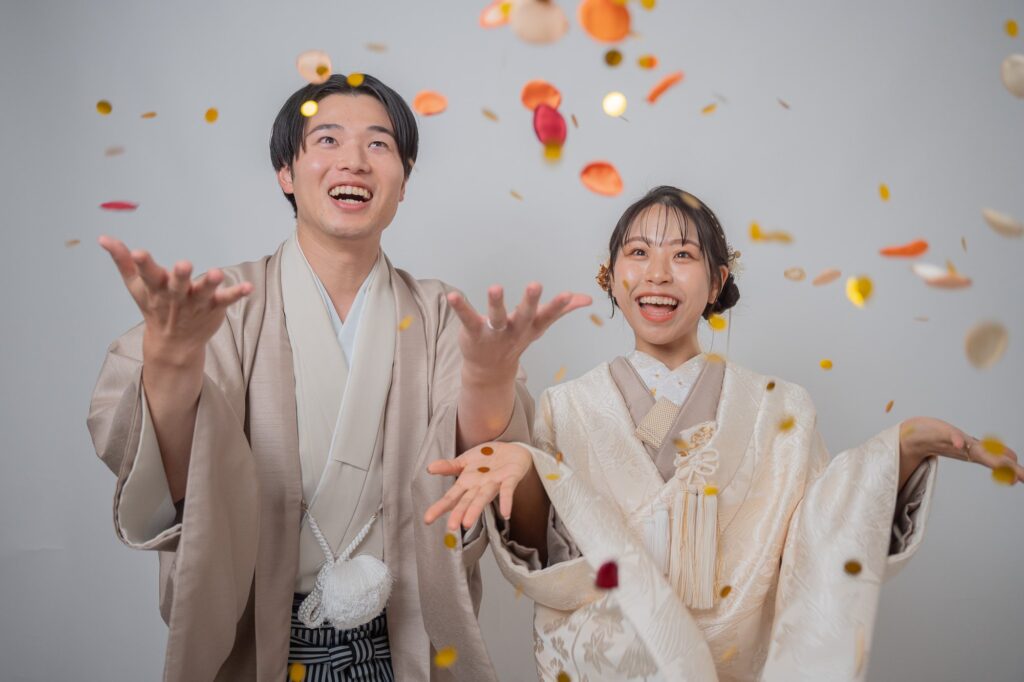 生成りの白無垢の花嫁と羽織袴の新郎がオレンジや金のコンフェッティを見上げて笑う和装前撮り