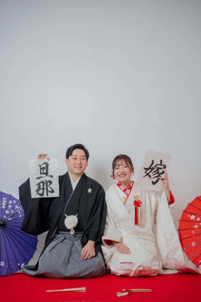 「旦那」「嫁」の習字アイテムを掲げて座る紋付袴の新郎と白無垢の花嫁