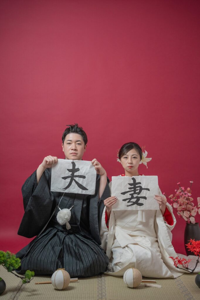 「夫」「妻」の習字の紙を持って畳の上に座る和装の新郎新婦