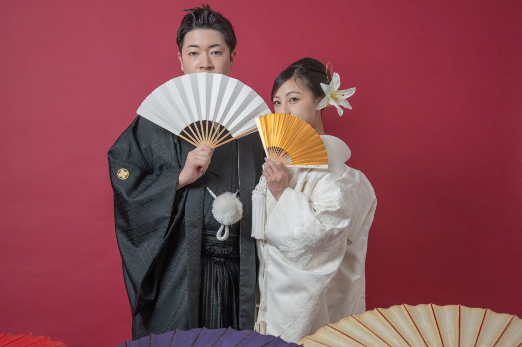 白と金の扇子を顔の横で広げる紋付袴の新郎と白無垢の花嫁