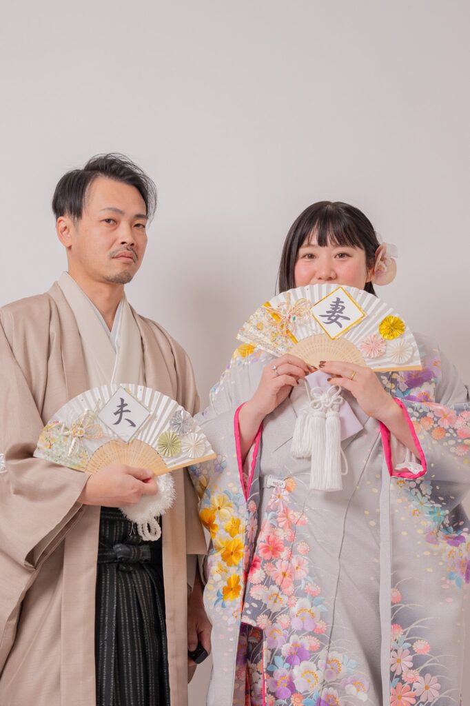 「夫」「妻」の文字入り扇子を持って並ぶ和装の新郎新婦の上半身ショット