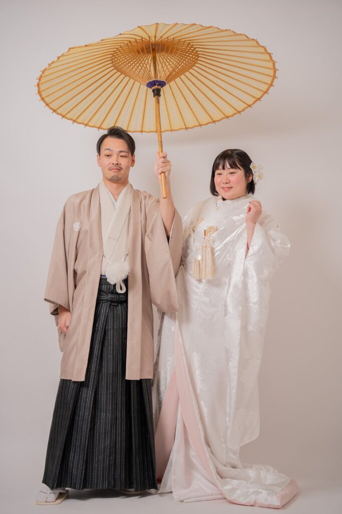 黄色の和傘をさした羽織袴の新郎と白無垢の花嫁が並ぶ全身ショット