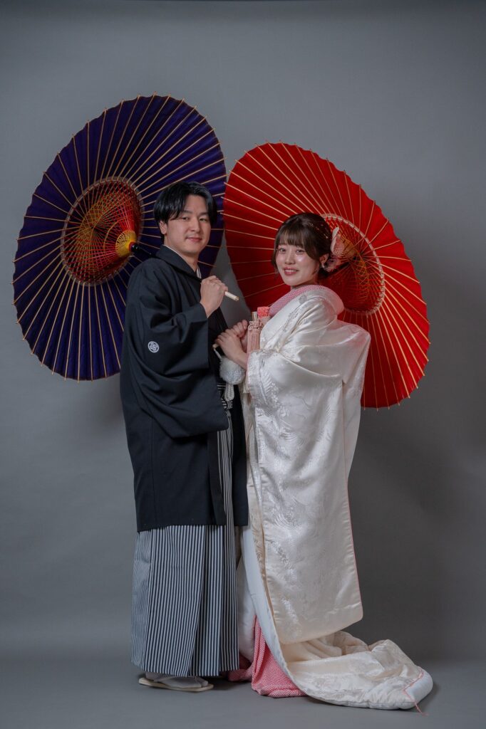 紫と赤の和傘を背に寄り添う白無垢の花嫁と紋付袴の新郎