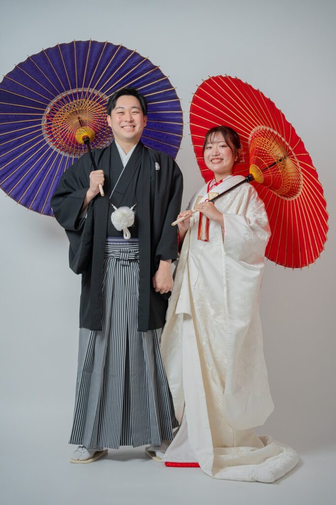 紫と赤の和傘を背に並ぶ白無垢の花嫁と紋付袴の新郎の全身ショット