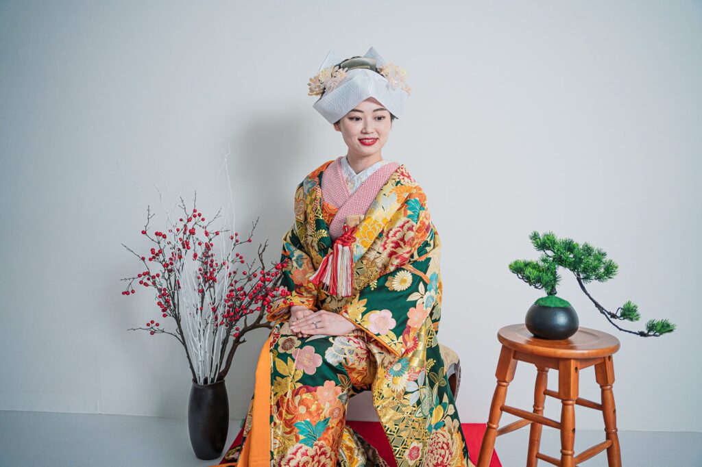 綿帽子をかぶった色打掛の花嫁が梅と盆栽のセットの前で座るスタジオ写真